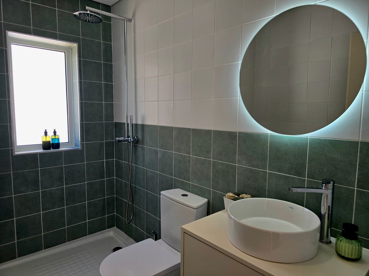 Bathroom with green tiles, a round lit mirror and a walk in shower